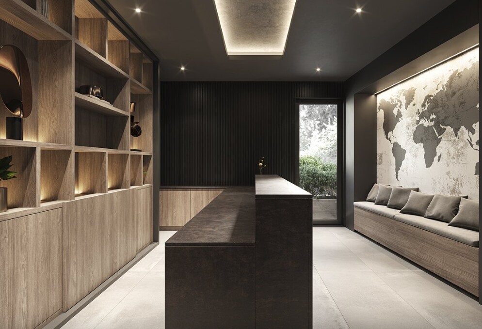 A modern reception area features sleek cabinetry and a dark countertop, illuminated by soft ceiling lights. A wall showcases a world map, while a window offers a view of greenery. A long bench with cushions provides seating, enhancing the space's welcoming design.