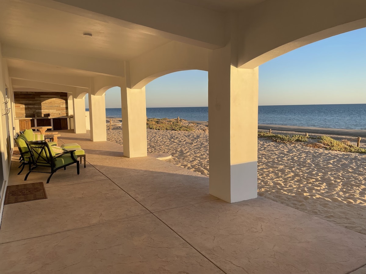 A spacious back patio is visible, featuring arched columns that create a welcoming open space. Comfortable patio chairs are arranged thoughtfully, with a view of the sandy beach and ocean beyond. The golden light of sunset enhances the tranquil atmosphere.
