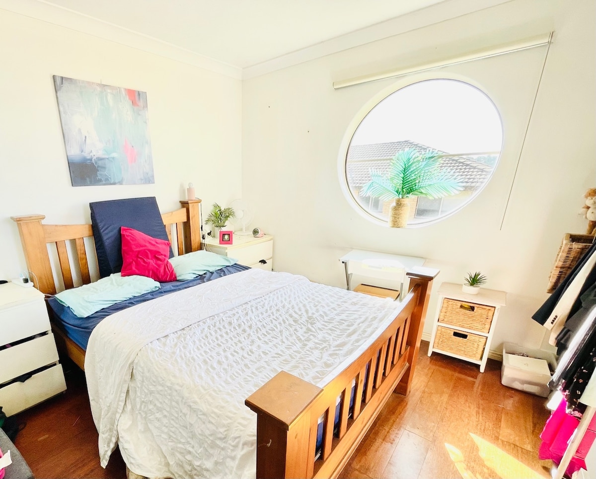 A serene bedroom features a wooden bed with a white quilt and colorful pillows. A round window allows natural light to fill the space, revealing a glimpse of greenery outside. A bedside table and a small desk enhance functionality, while a few decorative plants add freshness.