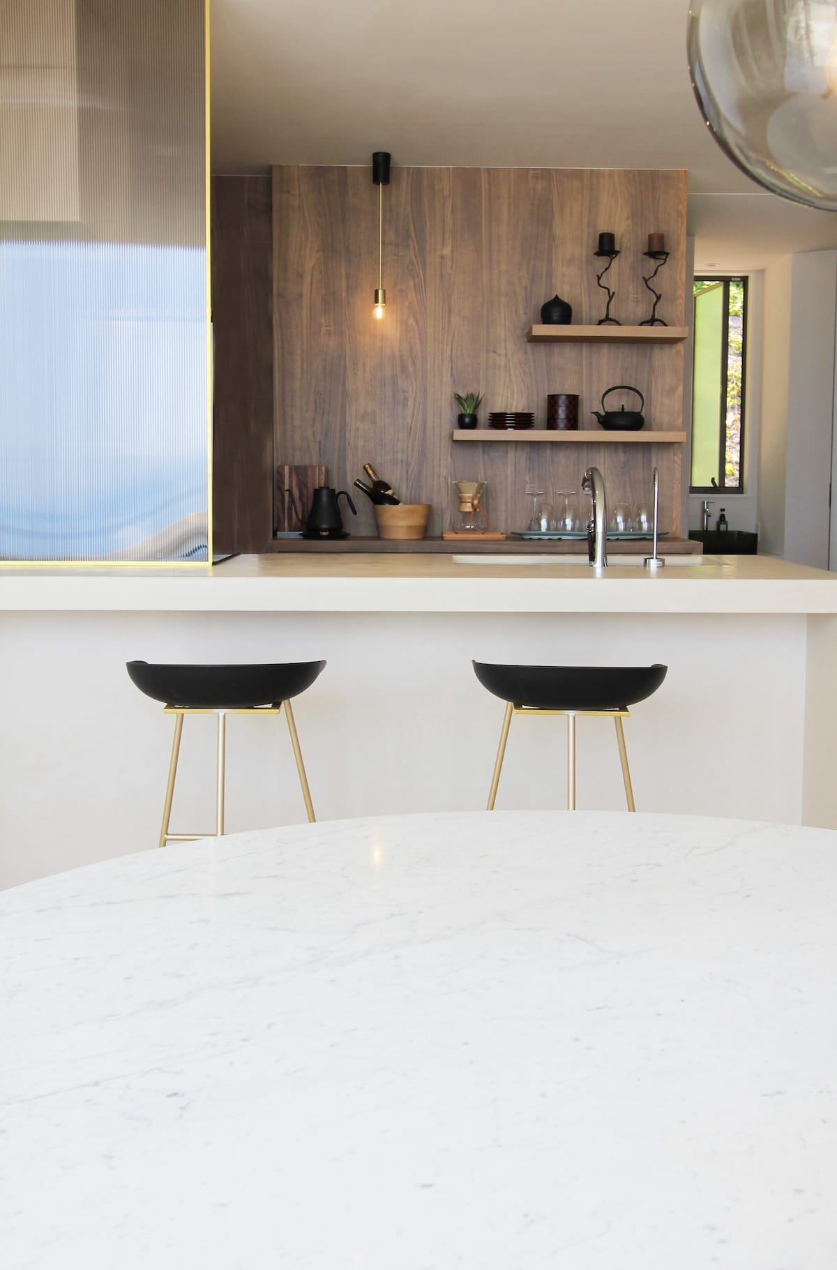 A modern kitchen space features a sleek marble-top island and two black bar stools with gold accents. The background showcases a minimalist wood panel with various kitchen utensils and decorative items, enhanced by soft lighting for a warm ambiance.