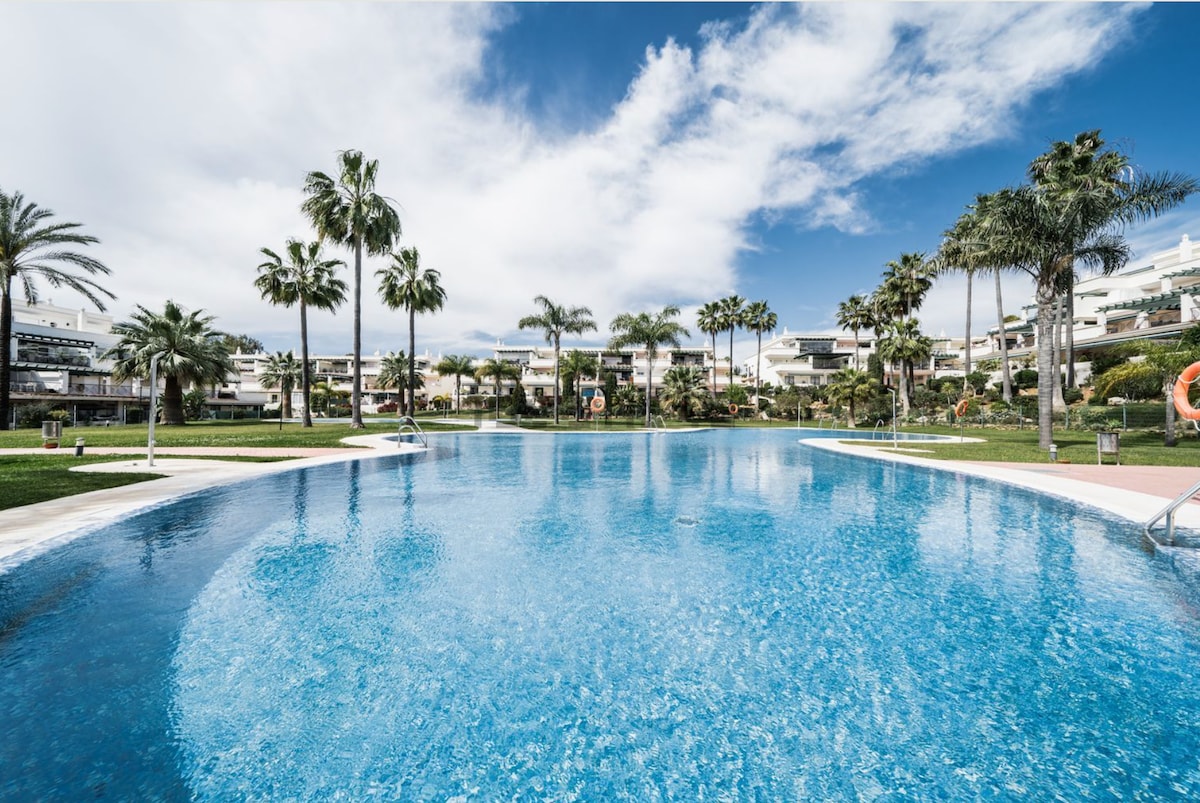 A large swimming pool is framed by well-manicured lawns and palm trees, offering a serene environment. The water is calm and clear, reflecting the blue sky and scattered clouds. Sun loungers and trees can be seen lining the pool area, enhancing the spacious atmosphere.