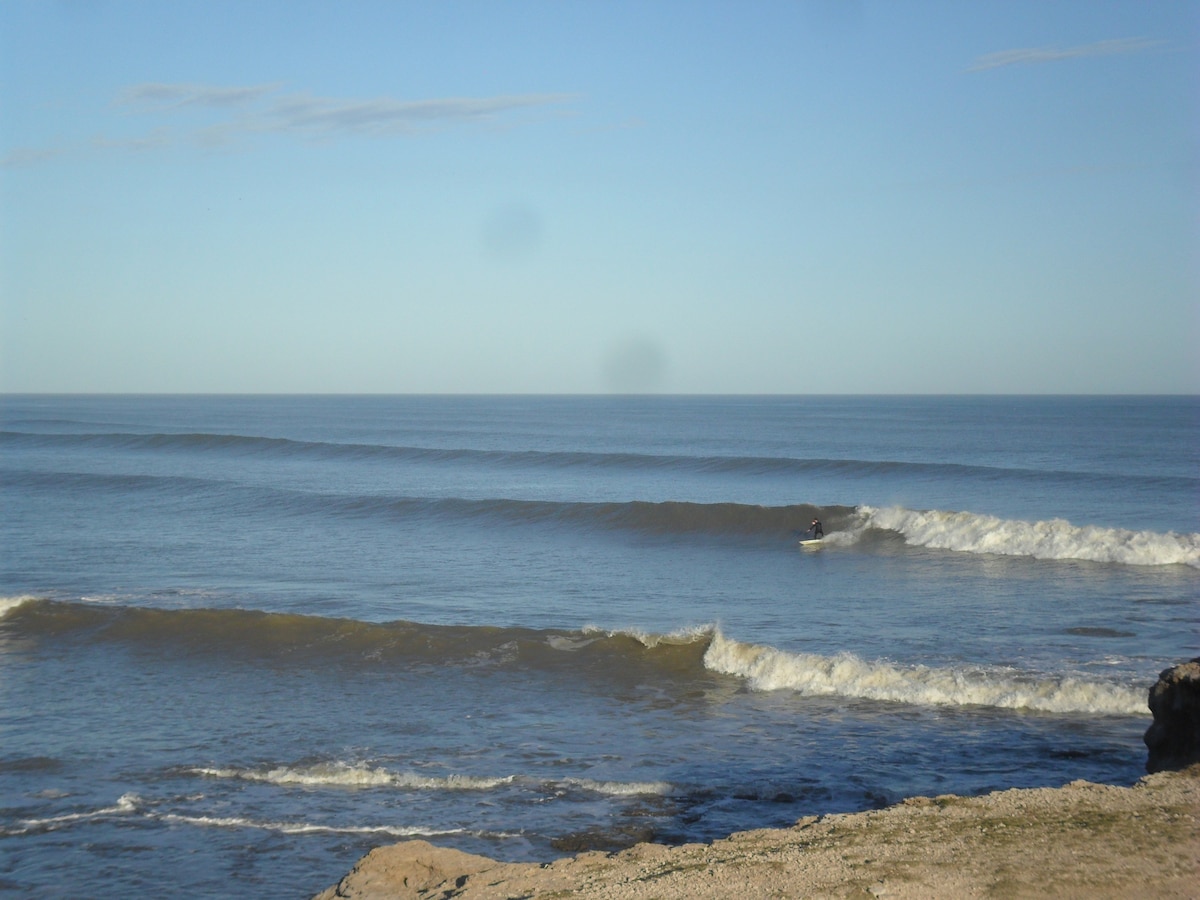 Waves gently roll toward the shore under a clear blue sky. A surfer is seen riding one of the waves, capturing the essence of seaside activities. The coastline appears rocky, providing texture against the tranquil ocean surface.