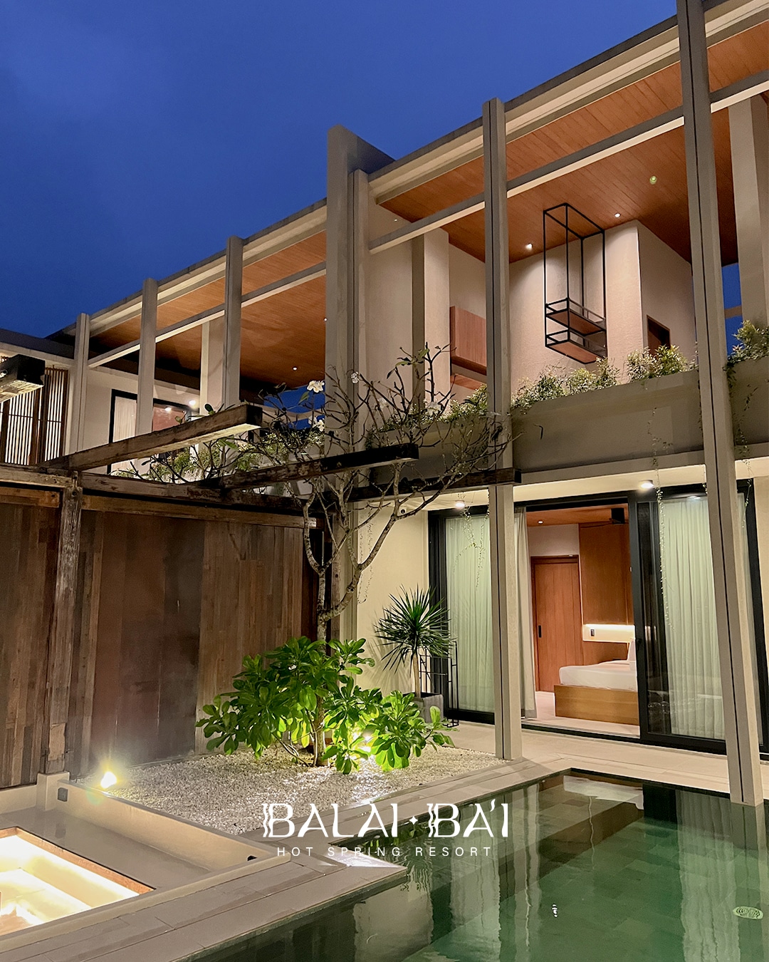The villa's exterior showcases a modern design, featuring wooden accents and expansive glass elements. A tranquil courtyard is adorned with greenery and a soothing pool reflects the evening sky. The upper levels are illuminated, highlighting the architectural details and inviting ambiance.