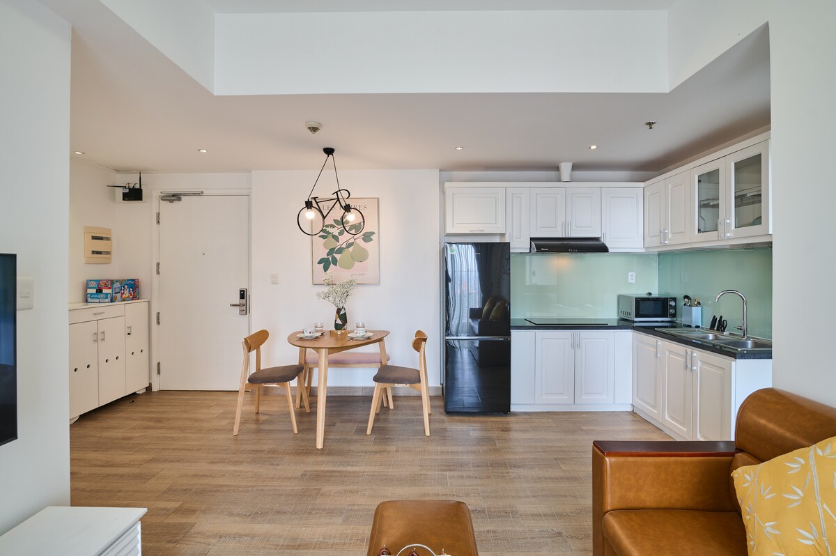 A modern dining area features a round wooden table with two chairs, positioned beside a sleek kitchen. The kitchen includes white cabinetry, a black refrigerator, and a minimalist glass backsplash. Soft natural light enters through a nearby window, illuminating the open space.