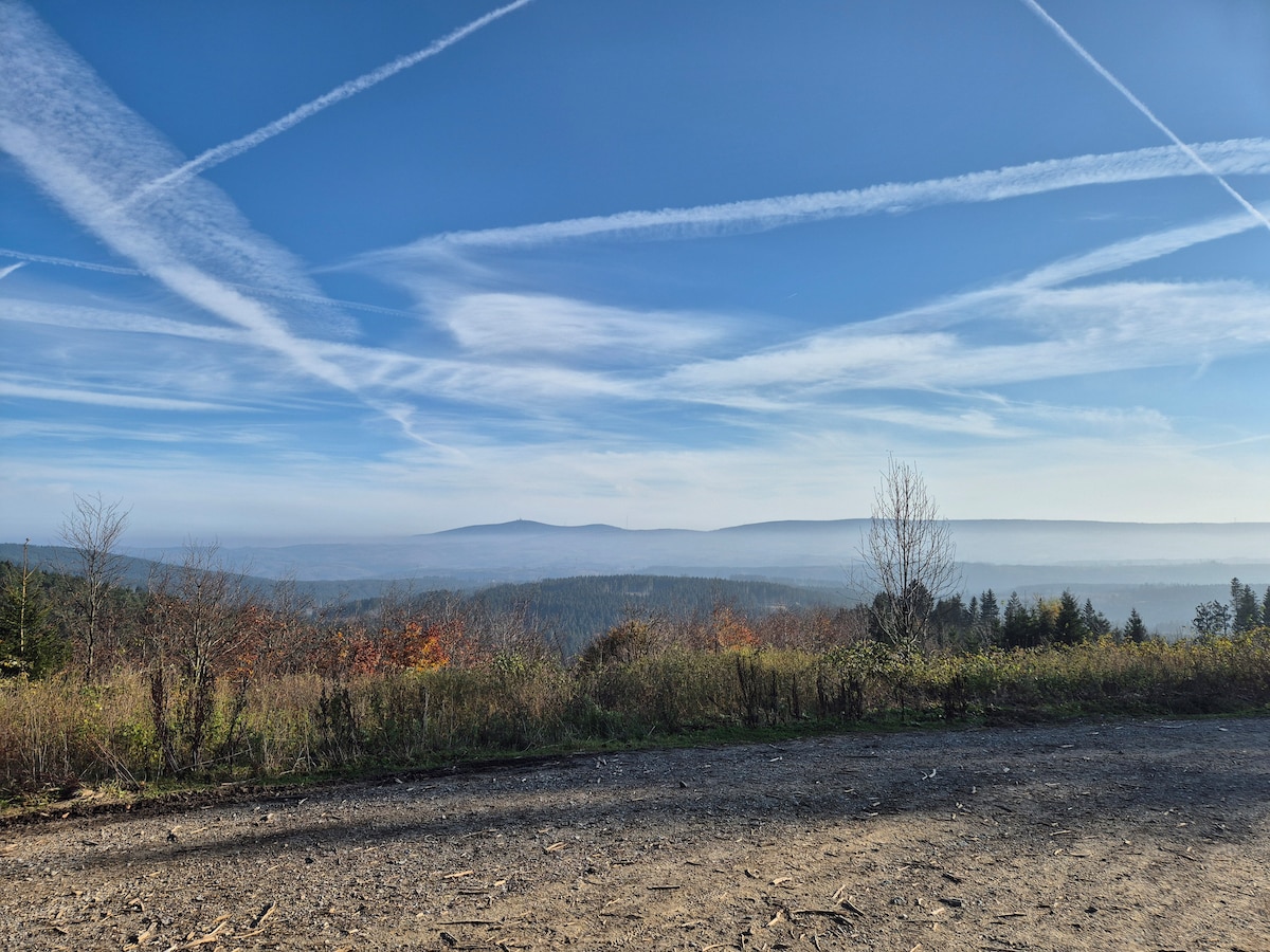 A serene landscape showcases rolling hills under a clear blue sky, with contrails forming distinctive patterns overhead. The foreground features a gravel path and patches of greenery, while the distant mountains provide a peaceful backdrop.