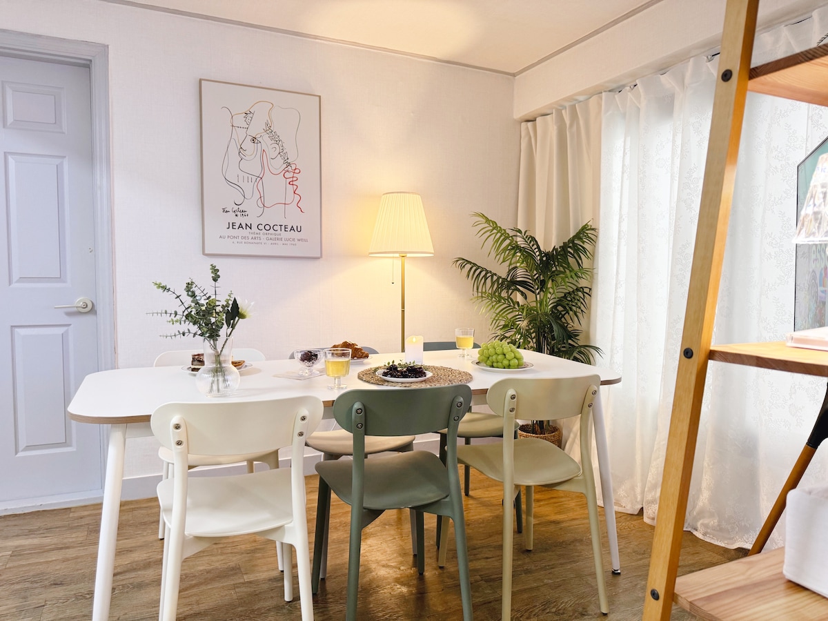 A dining area is featured, showcasing a white table surrounded by four chairs in soft colors. A decorative plant sits beside the table, and a warm lamp stands nearby, adding subtle illumination to the space. A wall art piece adds character to the area.