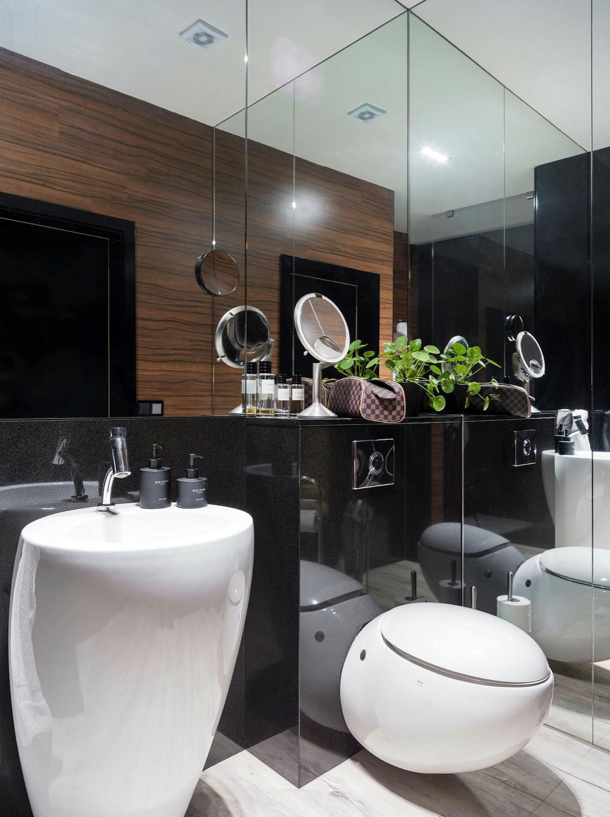 A modern bathroom is presented, featuring sleek black and brown wood paneling. The space includes a minimalist white sink and a rounded toilet. A circular mirror is mounted above the sink, reflecting the well-organized toiletries and a small green plant, enhancing the contemporary design.