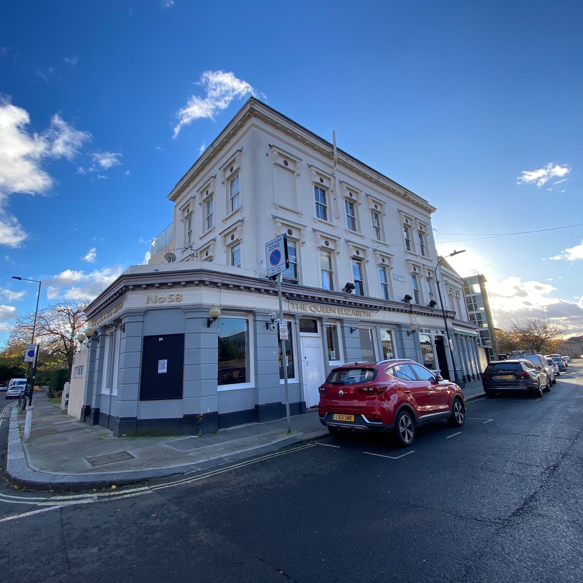 The exterior of the Queen Elizabeth hostel features a classic pub facade with large windows and a welcoming entrance. The structure is three stories high, painted in white, with decorative trim. A red car is parked nearby on the street, reflecting the urban environment.