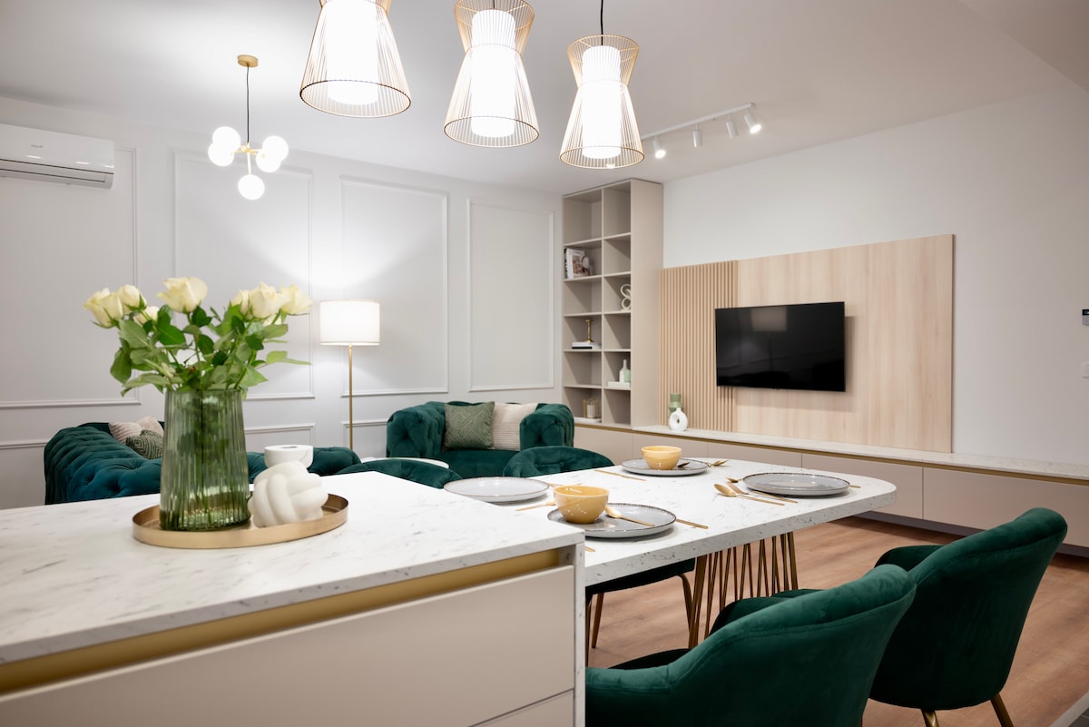 A stylish living area features a White marble countertop, with a vase of white roses taking center stage. A modern dining table with green chairs is set for a meal. Light fixtures hang from the ceiling, and a flat-screen TV is mounted on a wooden accent wall.