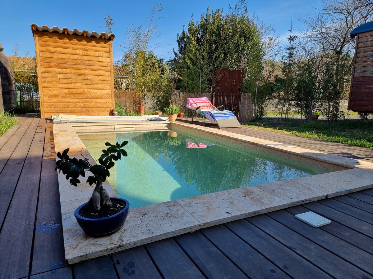 A serene swimming pool is bordered by natural stone, with wooden decking surrounding it. Chairs are positioned beside the pool for relaxation. Lush greenery and a wooden shed are visible in the background, completing the tranquil outdoor setting.