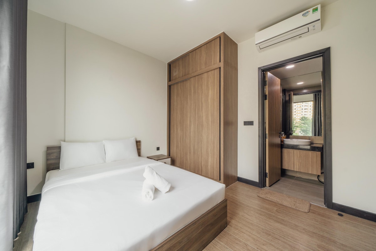 A modern bedroom features a spacious bed accompanied by a neatly rolled towel. Soft lighting enhances the simple decor, with a wardrobe and bedside table visible. A doorway leads to an en-suite bathroom, while large windows provide natural light.