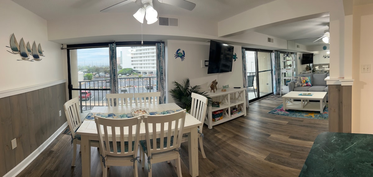 A spacious living and dining area is visible, featuring a large dining table surrounded by white chairs. Natural light flows through sliding glass doors leading to a balcony. Coastal decor accents the walls, and a television is mounted on the wall, enhancing the vibrant atmosphere.