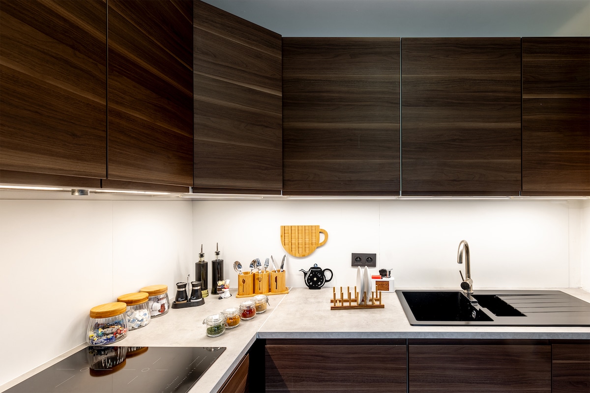 The kitchen features modern cabinetry with a sleek finish. A countertop is equipped with a stove and a stylish sink. Various kitchen utensils and containers filled with ingredients are displayed, providing an organized cooking environment. The lighting highlights the clean lines and functionality of the space.