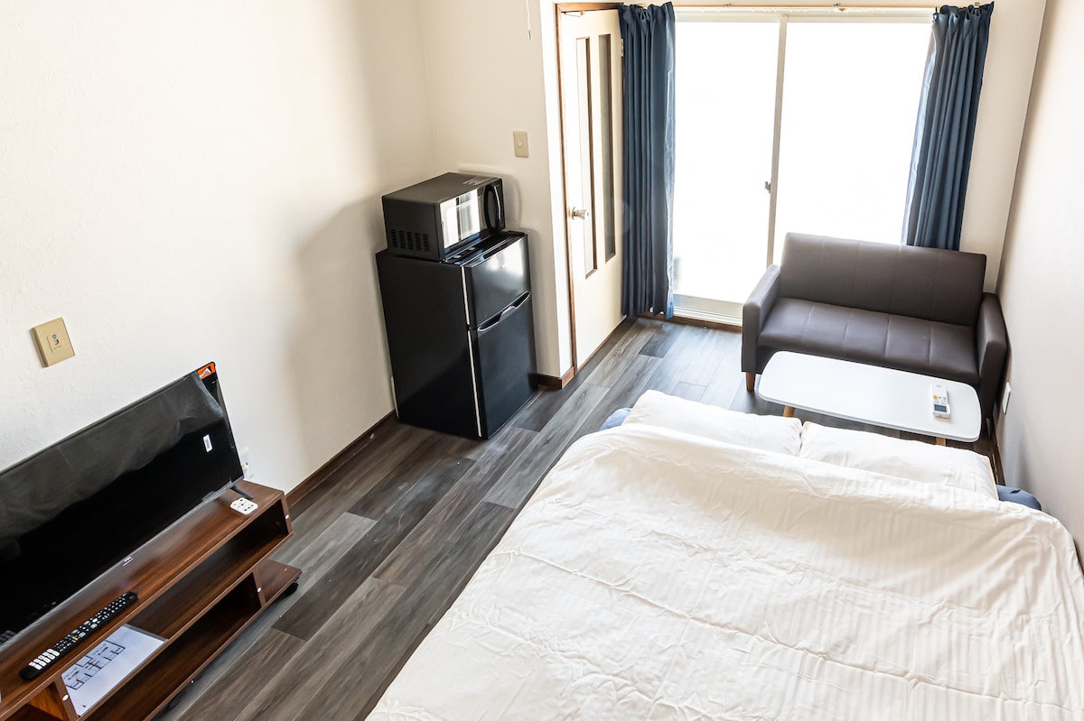 A bright and simple room features a spacious bed with crisp linens, a sleek couch, and a small table. A modern kitchenette includes a microwave and refrigerator. Light is allowed in through the large window, with dark curtains framing the view.