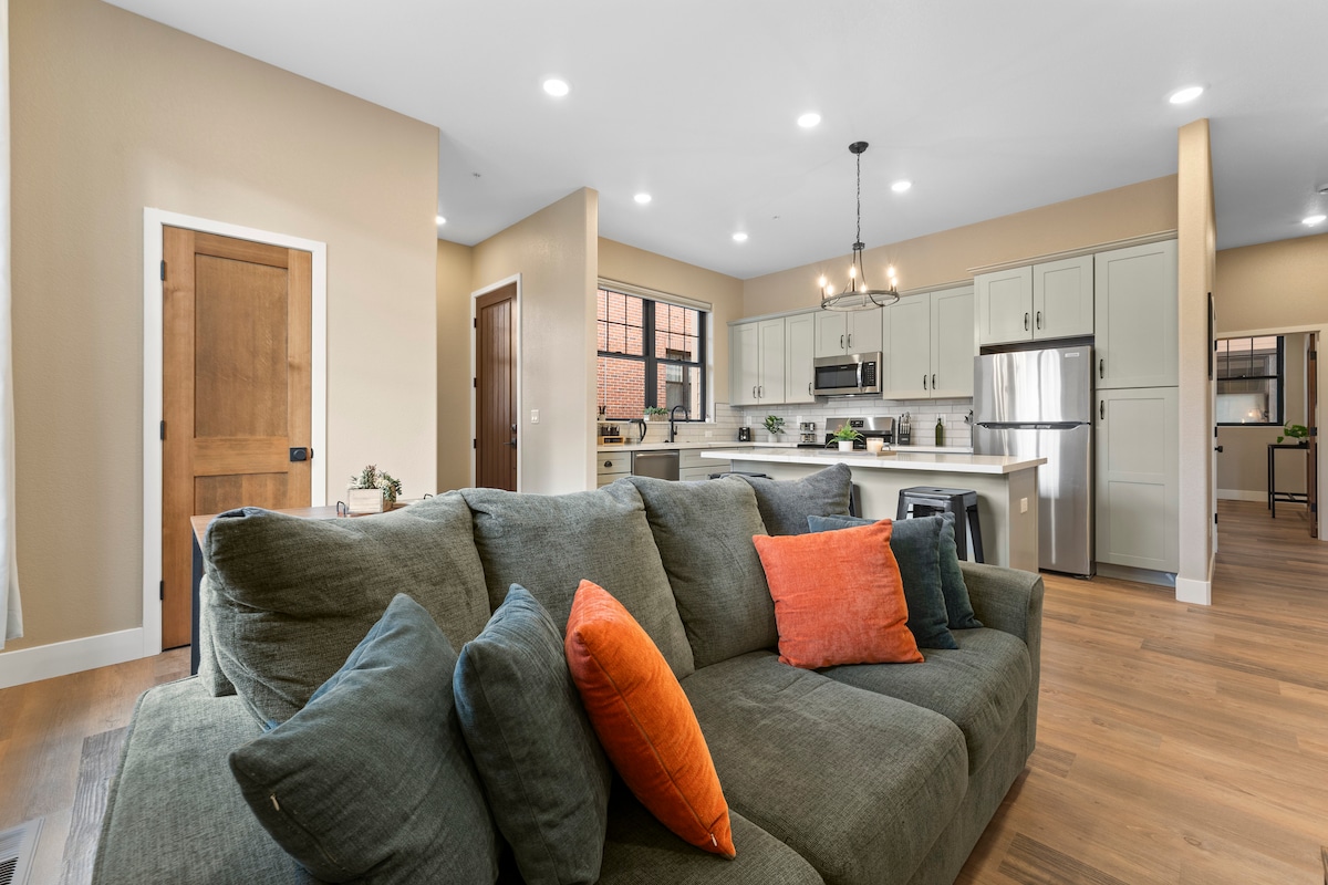 A spacious living area features a comfortable gray sofa adorned with colorful throw pillows. An open kitchen, equipped with modern appliances and fixtures, is visible in the background. Warm, neutral tones fill the room, complemented by natural light from the windows.