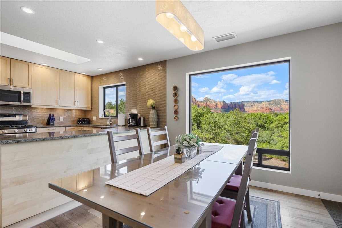 A modern kitchen and dining area features warm wooden cabinetry and stainless steel appliances. A large window offers expansive views of the surrounding mountains, while a dining table with multiple chairs provides ample seating. Bright and natural light fills the space.