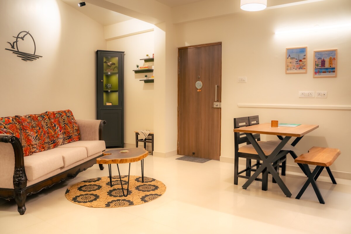 A spacious living area features a patterned sofa and a wooden coffee table at its center. A dining table with wooden benches is positioned to one side. Wall art adds a touch of color, and a doorway leads to the entrance.