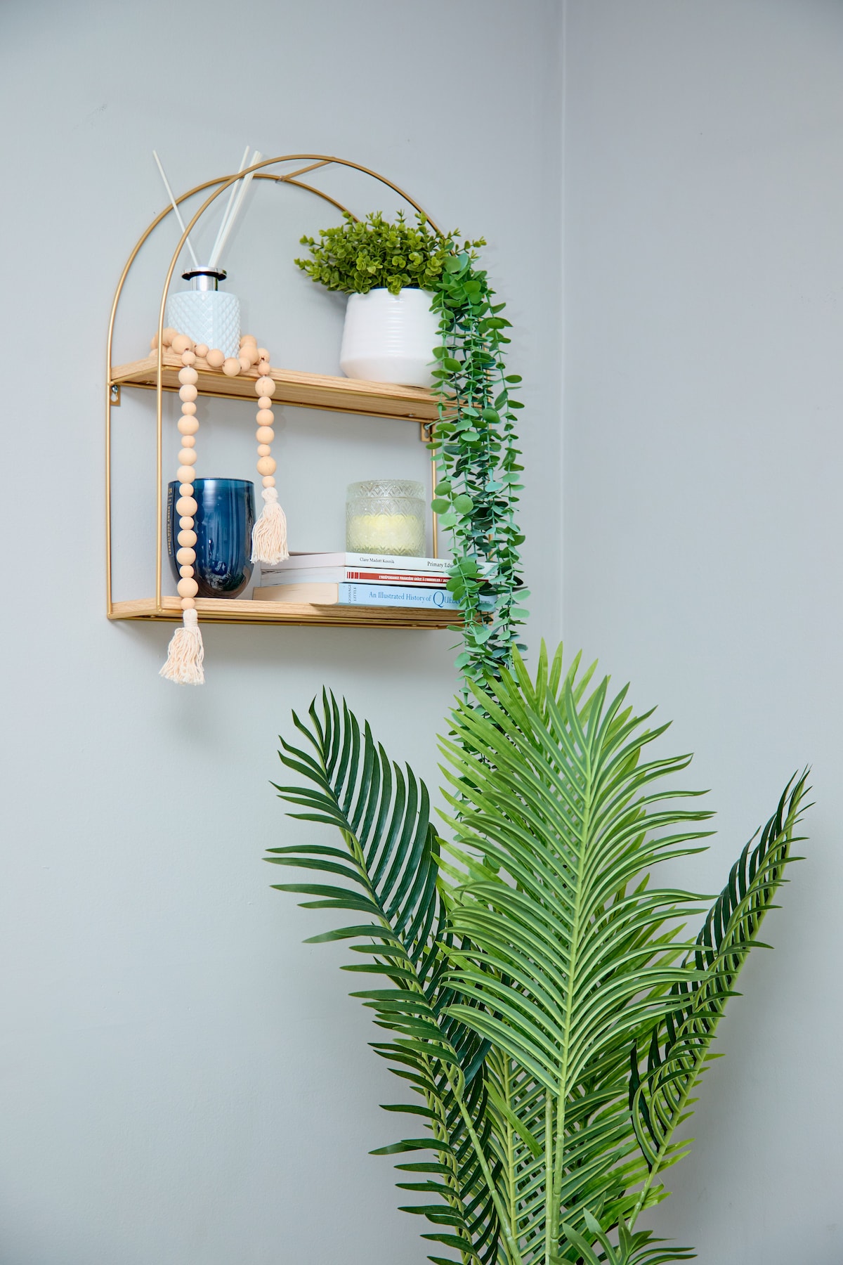 A wooden shelf is mounted on a light gray wall, featuring decor items such as a potted plant, a blue glass vase, and a white ceramic container. Greenery trails down from the shelf, complemented by a tall, leafy plant positioned nearby.