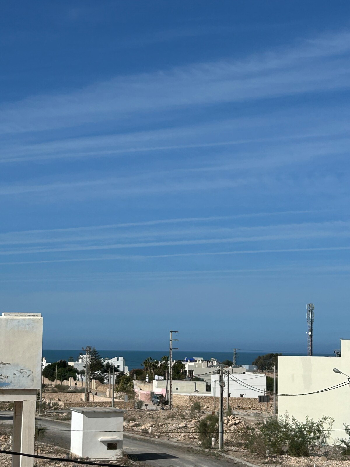 A distant view of the sea is visible under a clear blue sky, with wispy clouds stretching across the horizon. In the foreground, a blend of residential buildings and open land is observed, capturing the coastal setting of the area.
