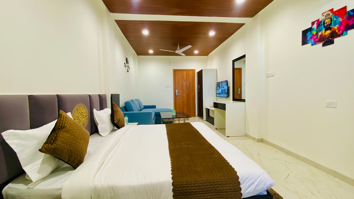 A well-lit room features a spacious bed adorned with decorative pillows. A sleek, blue couch provides additional seating across from a flat-screen television mounted on a white cabinet. Light-colored walls and a dark wood ceiling contribute to a modern aesthetic.