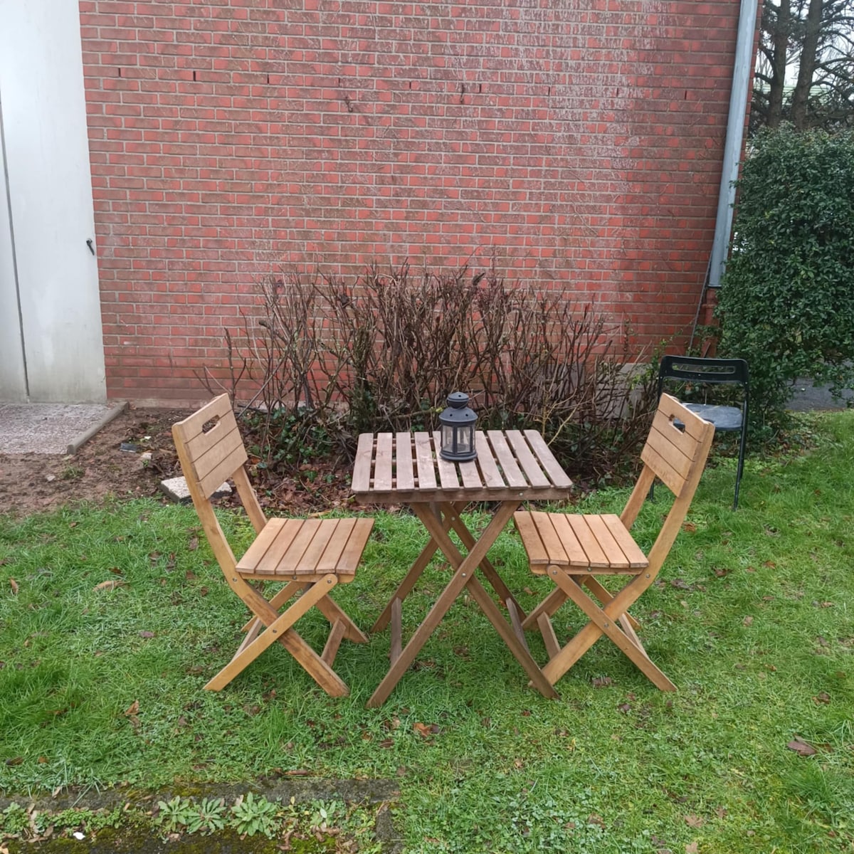 A small outdoor terrace features a round wooden table with two matching folding chairs. A lantern is positioned on the table, surrounded by a grassy area and low shrubbery against a red brick wall.