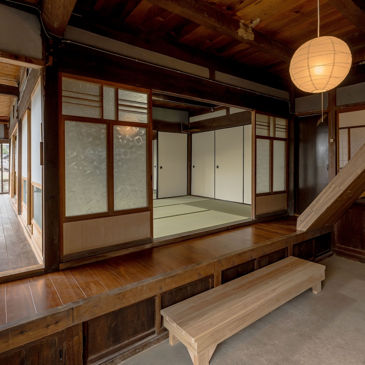 A wooden entrance area features a low bench and traditional decor. The adjacent room displays sliding doors and tatami mat flooring, creating a calm and spacious atmosphere. Natural light enhances the warm wooden tones throughout the space, connecting indoor and outdoor environments.