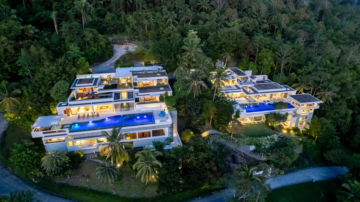 An aerial view showcases two adjacent villas nestled in lush, tropical greenery. The structures feature multiple levels with large terraces and inviting pools reflecting the evening light. Paved driveways connect the villas, enhancing accessibility among the beautifully landscaped surroundings.