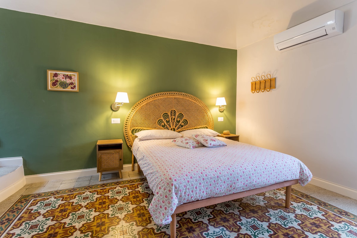 A spacious sleeping area features a large bed with a patterned duvet cover. Two bedside lamps provide soft illumination, and a decorative wall hangs above the bed. A colorful area rug lies beneath, while a slight texture of the green wall enhances the room's character.