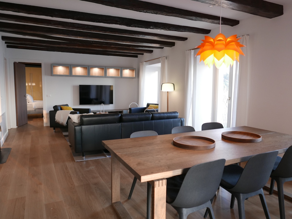 An open floor plan is displayed, featuring a dining area with a wooden table and gray chairs. A spacious seating arrangement with dark furniture is seen, accompanied by a TV mounted on the wall. Natural light enters through large windows, enhancing the modern decor.