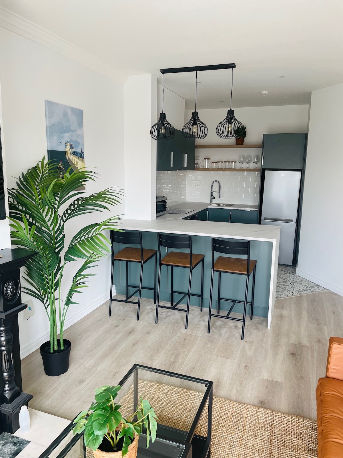 A modern kitchen area features sleek cabinetry and a light countertop, complemented by three high chairs at the bar. Decorative lighting hangs above, while a potted plant adds a touch of greenery. The space transitions seamlessly into the adjacent cozy sitting area.