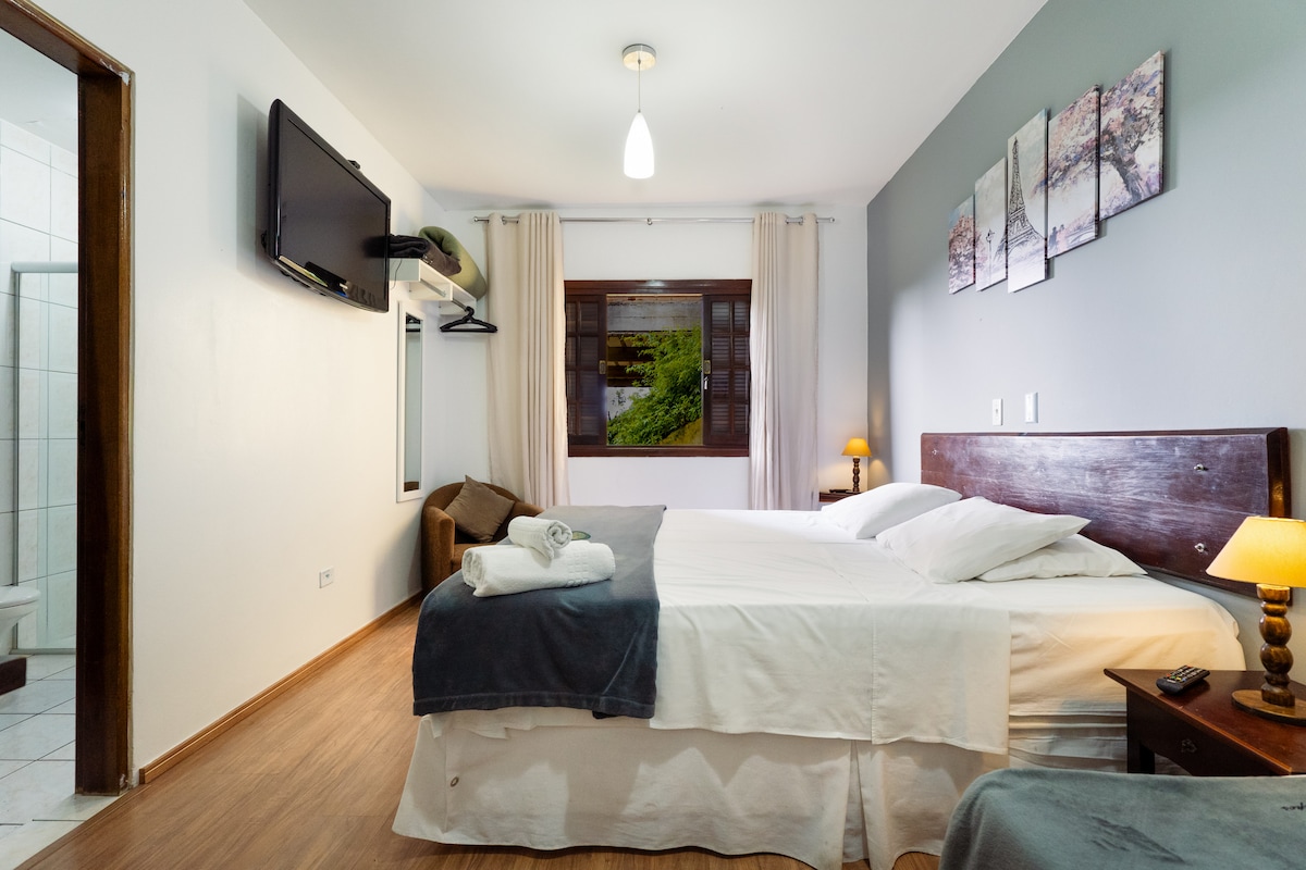 A well-lit room features a spacious bed with crisp white linens, accented by a soft gray throw. A flat-screen TV is mounted on the wall, and a cozy seating area is visible. Natural light filters through a window, complemented by serene wall art.