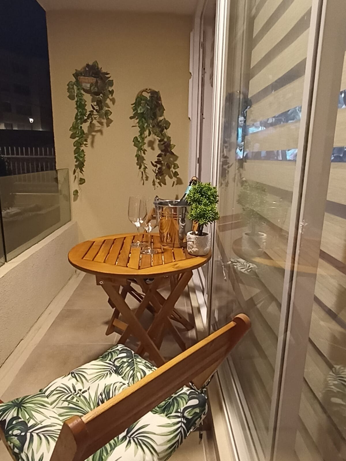 A small balcony space is depicted, featuring a wooden folding table set for two. A pair of glasses sits atop the table, accompanied by a bottle. Greenery is seen hanging on the wall, while a chair with a patterned cushion complements the inviting setting.