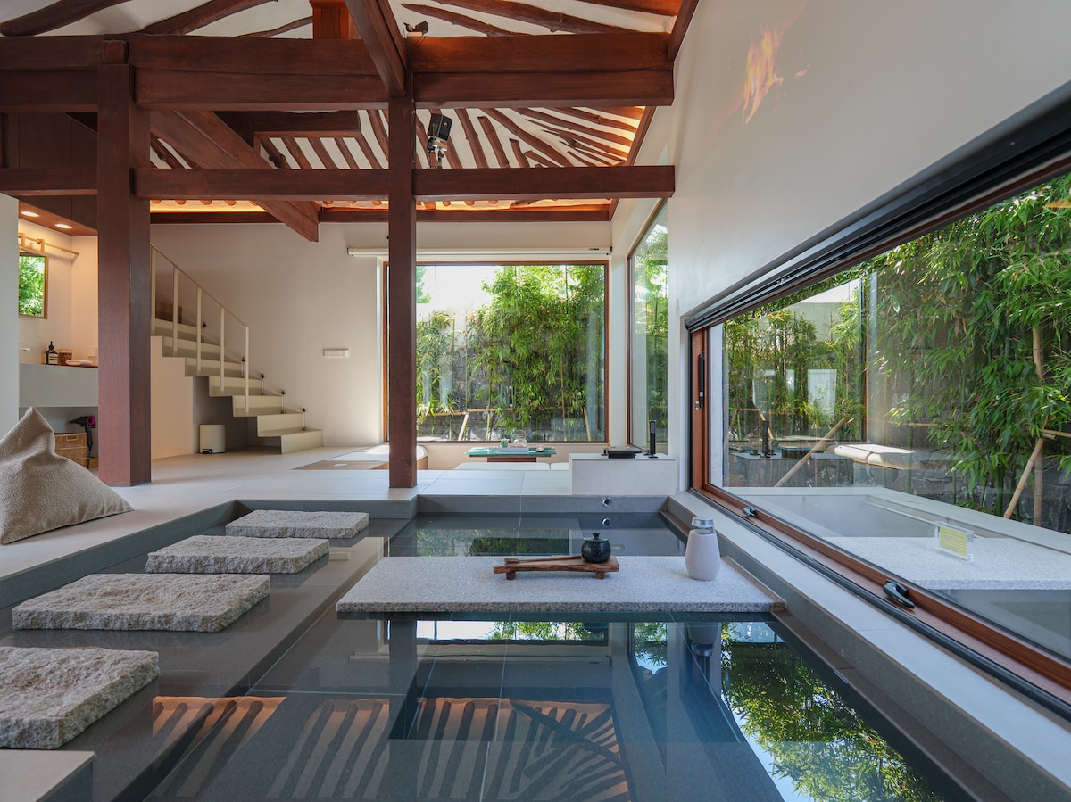 A serene interior space features large windows allowing natural light to illuminate the area. A tranquil indoor pool is complemented by stone stepping stones, while bamboo plants are visible outside, creating a calming connection to nature.