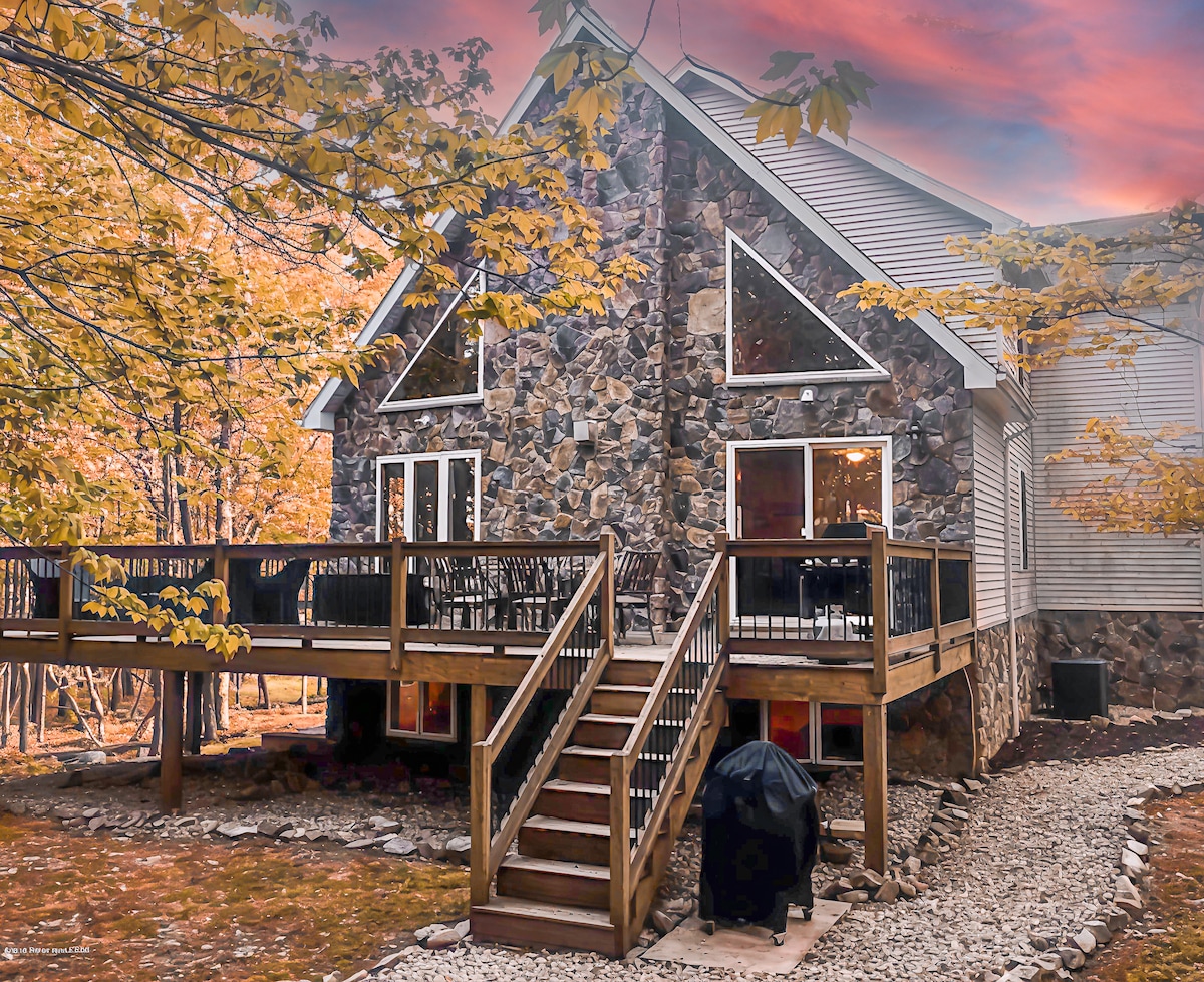 The exterior of a spacious stone house is showcased, featuring multiple large windows and a wraparound wooden deck. A set of stairs leads up to the entrance, while a grill is positioned on the deck, surrounded by autumn foliage.