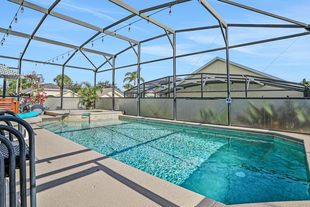 A spacious, screened pool area features a clear water pool and attached spillover spa. Surrounding areas are adorned with greenery and hanging lights, providing a relaxed ambiance. The pool deck is designed for lounging with patio furniture positioned nearby.