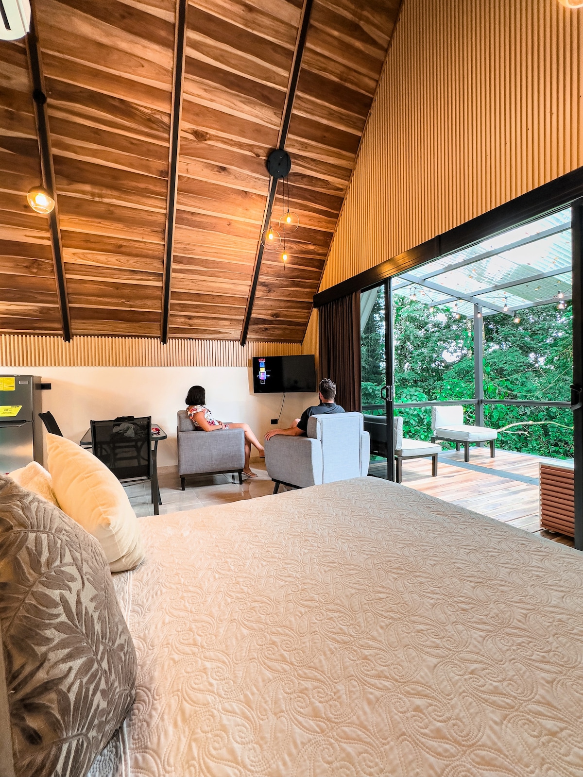 A spacious interior features a cozy seating area with light grey armchairs positioned toward a mounted television. A comfortable bed is visible in the foreground, while large glass doors open to a view of lush greenery. Warm wooden beams accent the high ceiling.