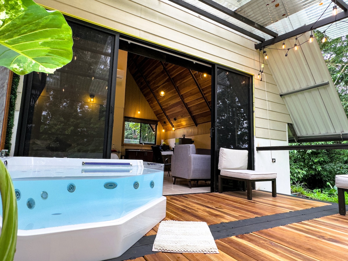 A private outdoor jacuzzi is positioned beside a wooden deck, inviting relaxation. Large glass doors lead into a spacious interior, showcasing a cozy living area. Lush greenery surrounds the space, enhancing the connection with nature.