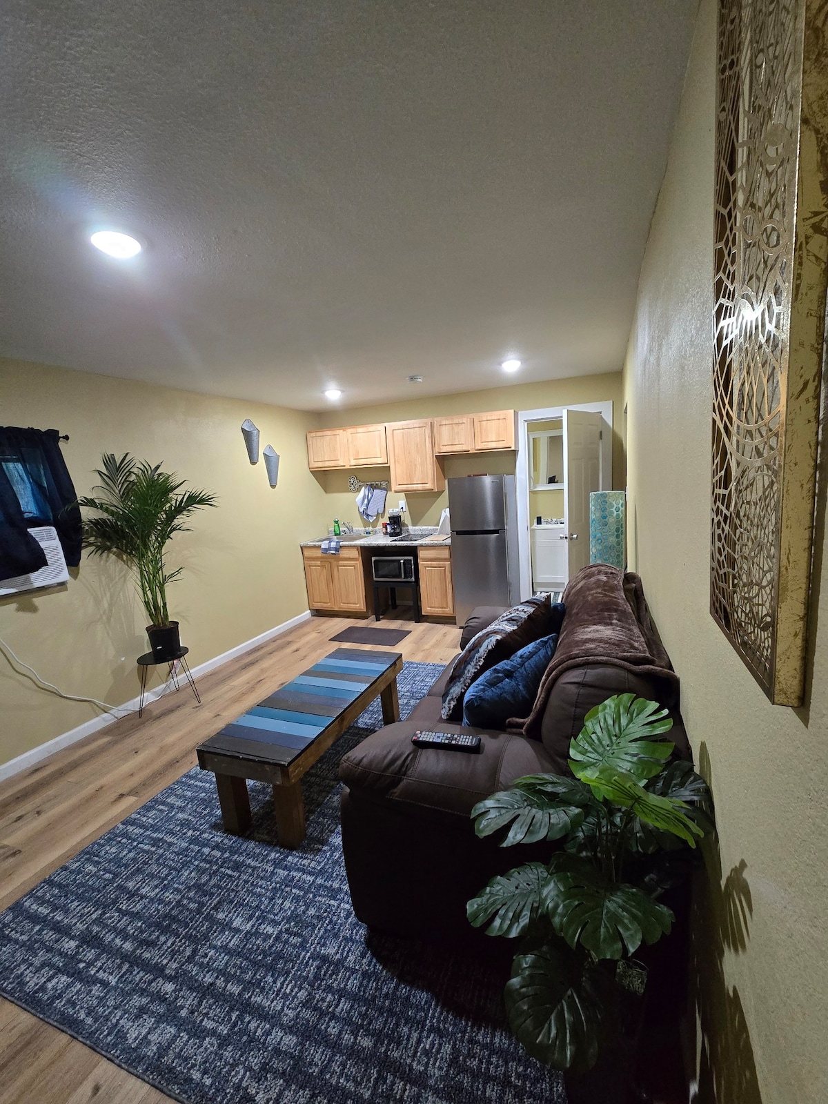 A comfortable living area features a dark brown couch with decorative pillows, situated on a textured blue rug. A wooden coffee table sits in front, and a kitchenette is visible in the background, showcasing light wood cabinets and modern appliances. A potted plant adds a touch of greenery.