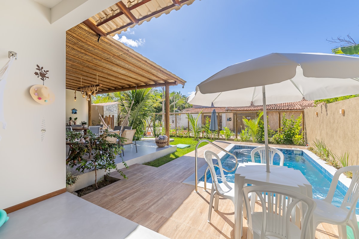 An outdoor area features a swimming pool surrounded by greenery, with a shaded patio including a thatched roof. Seating arrangements with tables and chairs are present under the patio, providing a serene environment. Sunlight illuminates the space, enhancing the inviting atmosphere.