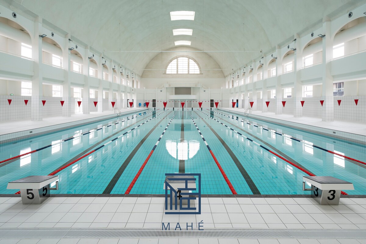 A spacious indoor swimming pool is showcased, featuring multiple lanes marked by floating lane dividers. The bright, airy environment is illuminated by natural light filtering through tall windows, while red triangular flags hang above each lane, creating a vibrant and inviting swimming atmosphere.