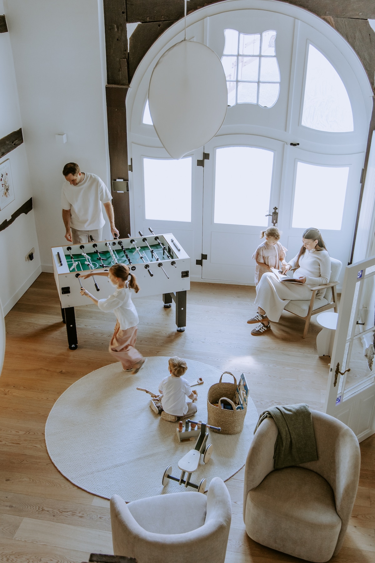 A spacious living area features a foosball table occupied by an adult, while children play on the floor. Natural light streams through large windows, illuminating the wood floor. Comfortable chairs are arranged nearby, creating a welcoming space for family gatherings.