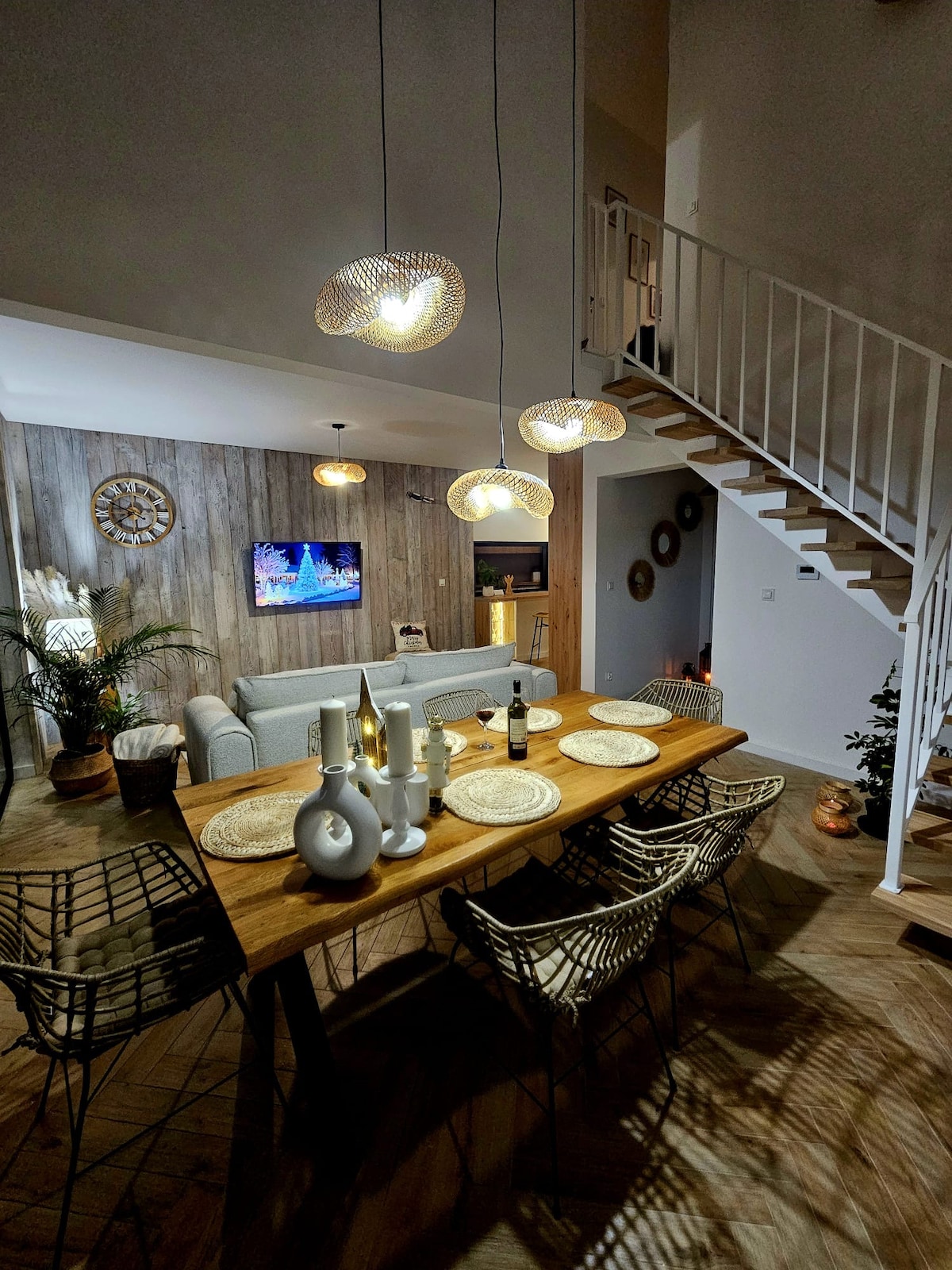 An inviting dining area features a wooden table set for six, accompanied by stylish wire chairs. Soft lighting from woven pendant lamps illuminates the space, highlighting a cozy sofa nearby. A staircase leads to the upper level, creating an open and airy atmosphere.
