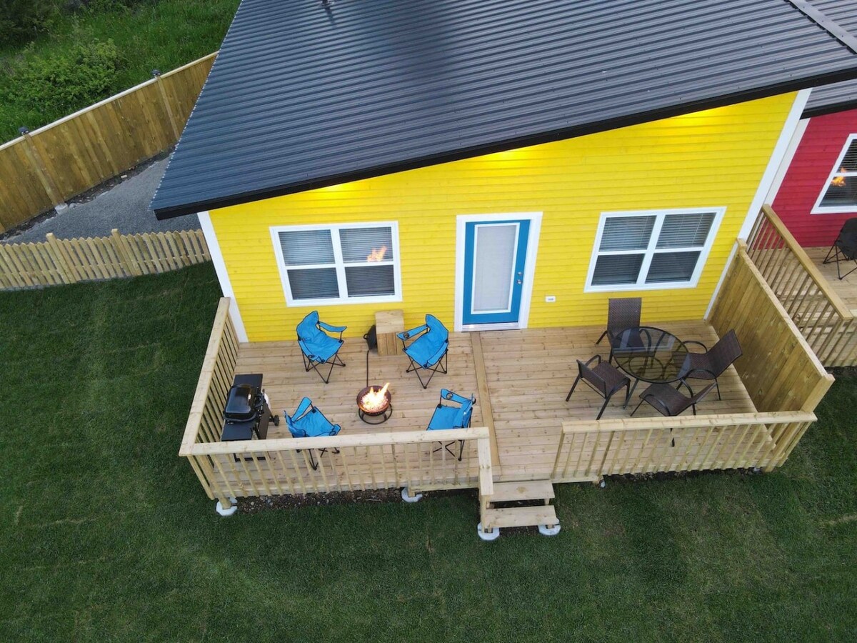 A vibrant yellow house is shown from above, featuring a spacious wooden deck. Blue outdoor chairs are arranged around a fire pit, creating a cozy gathering spot. A table and additional seating are positioned near the entrance, with green grass surrounding the area.