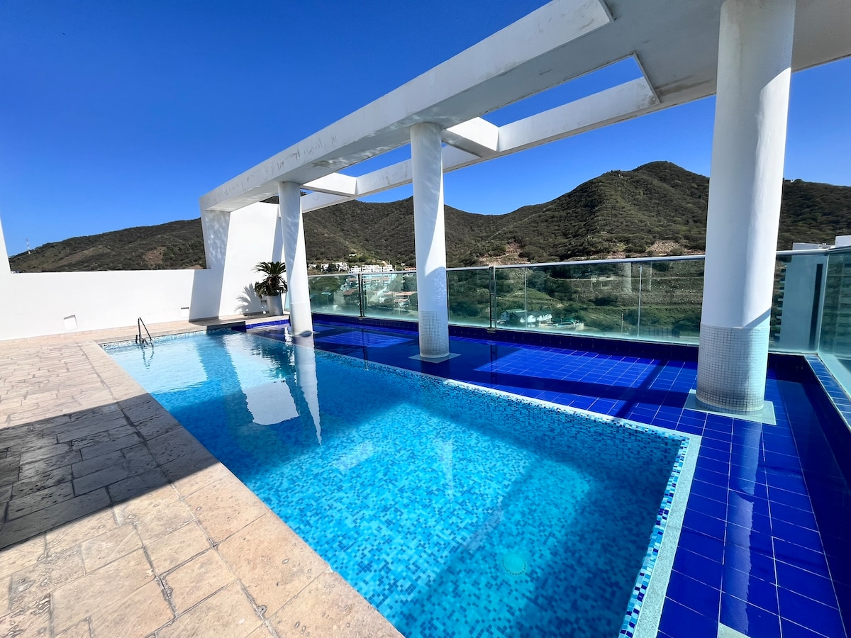 A rooftop pool is framed by modern architecture, featuring clear blue water surrounded by tiled decking. Lush greenery is present on one side, and distant mountains provide a scenic backdrop under a clear blue sky. Glass railings enhance the open feel of the space.