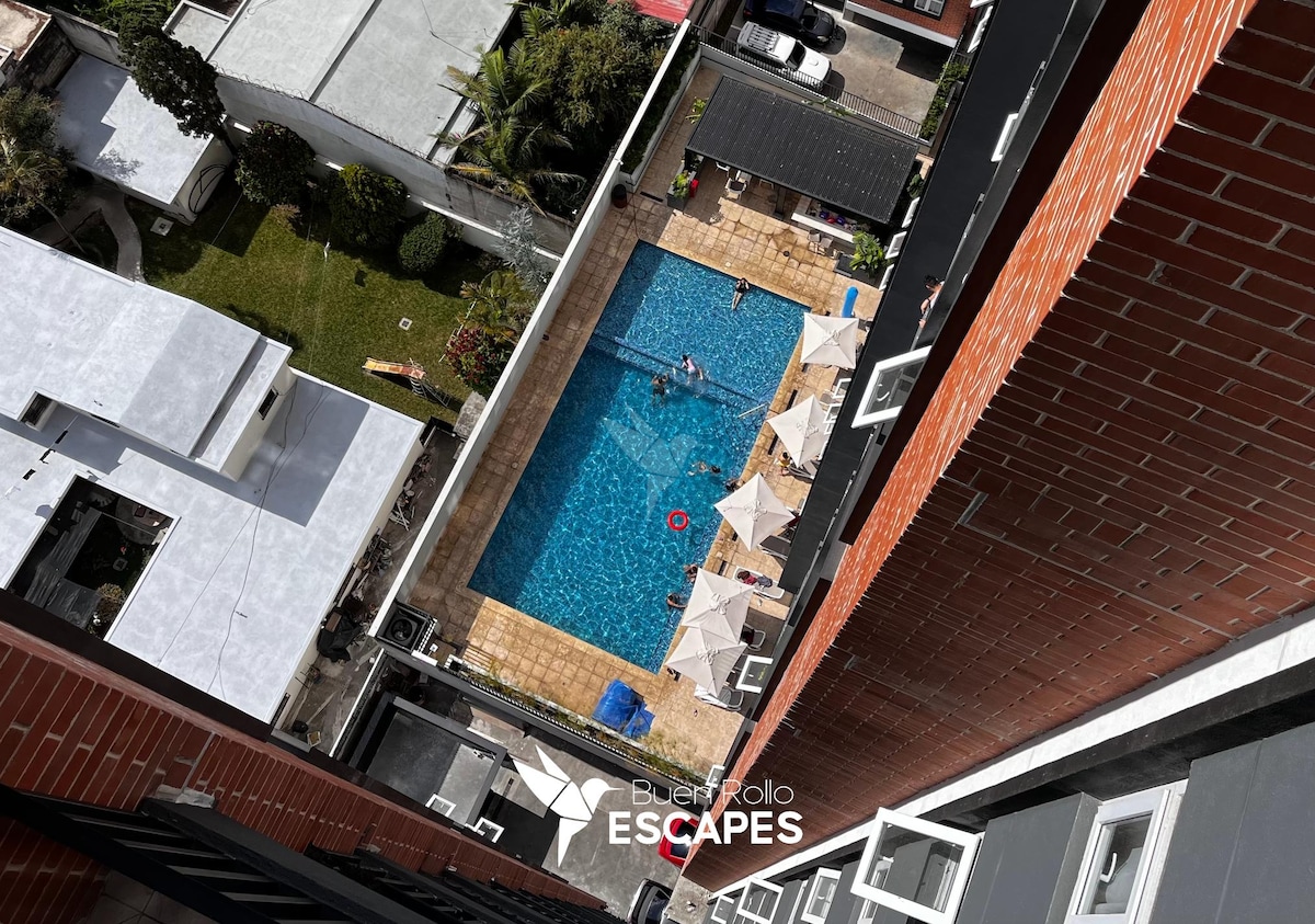 An aerial view of a spacious outdoor pool area is captured, featuring multiple sun umbrellas around the water. A patio with lounge chairs is visible, surrounded by lush greenery and well-maintained landscaping, creating an inviting atmosphere for relaxation.