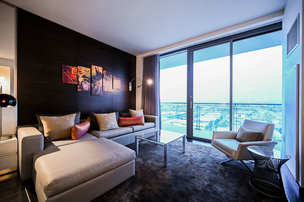 A modern living area features a sectional sofa adorned with decorative pillows, positioned beside a glass coffee table. Large floor-to-ceiling windows offer expansive views of the landscape, while a minimalist chair complements the contemporary design.