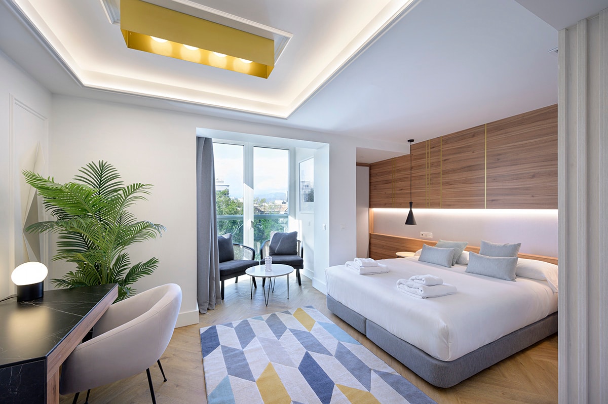 A modern bedroom features a spacious bed with neatly arranged linens and multiple pillows. A seating area includes two chairs and a small table. Natural light fills the room through large windows, and a plant adds a touch of greenery to the corner.