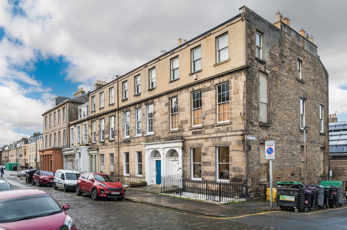 The exterior of a Georgian building is presented, showcasing a classic stone façade. Large windows and a white main entrance are visible. A quiet street is lined with parked vehicles and bins, complemented by a partly cloudy sky in the background.