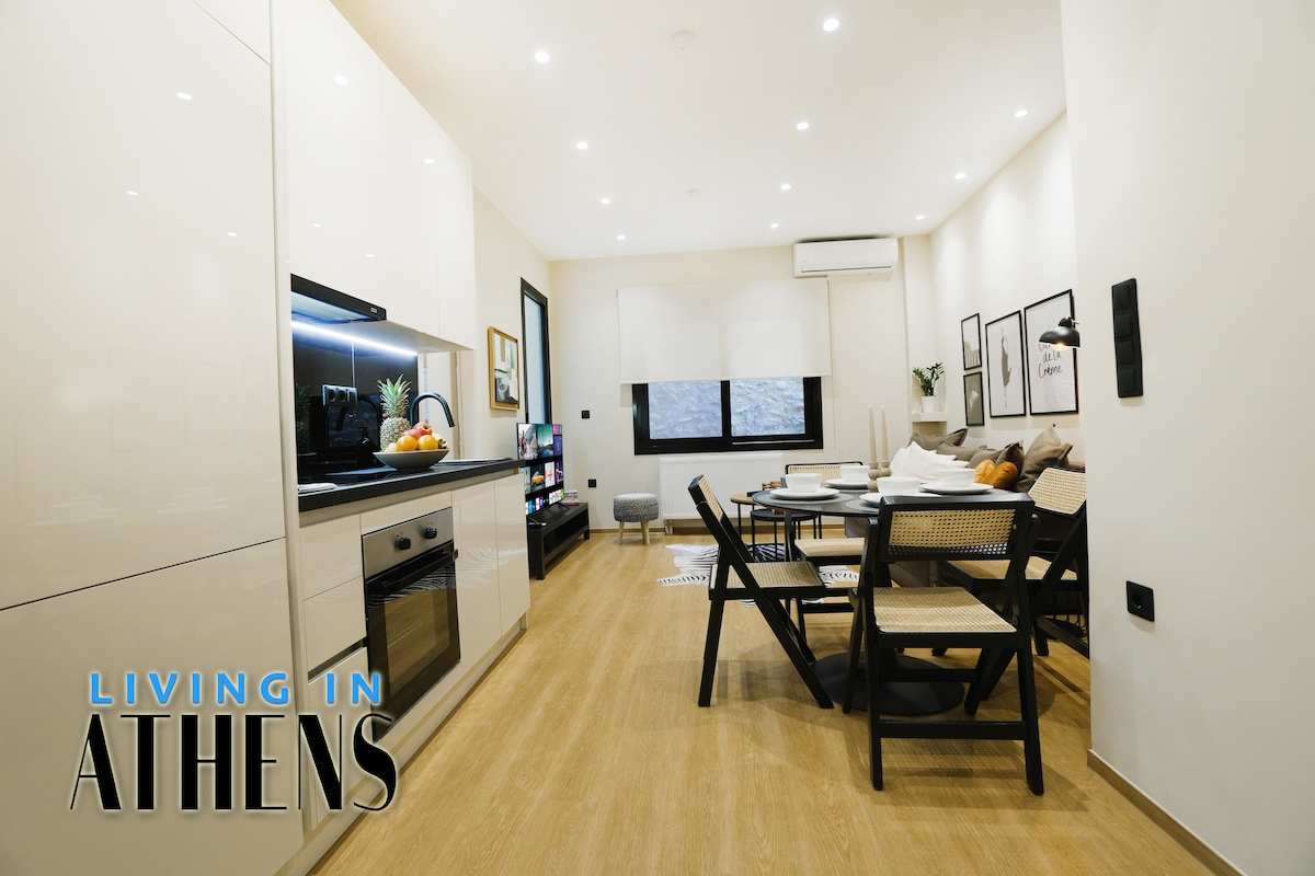 A modern kitchen and dining area is depicted, featuring sleek white cabinetry and stainless-steel appliances. A wooden dining table accommodates four chairs. The living space includes a multi-functional area with a cozy sofa and entertainment unit, illuminated by natural light through the windows.
