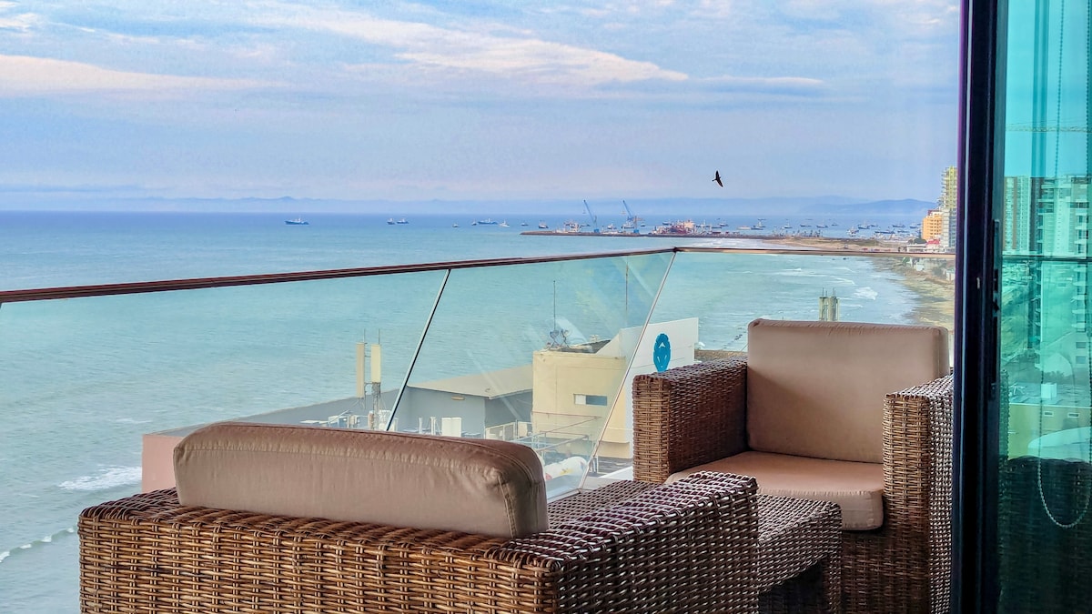 A balcony area is shown, featuring two wicker armchairs with plush cushions. A glass railing offers unobstructed views of the sea and coastline, creating a serene environment. The horizon displays a soft blend of blue tones, with boats visible on the water.